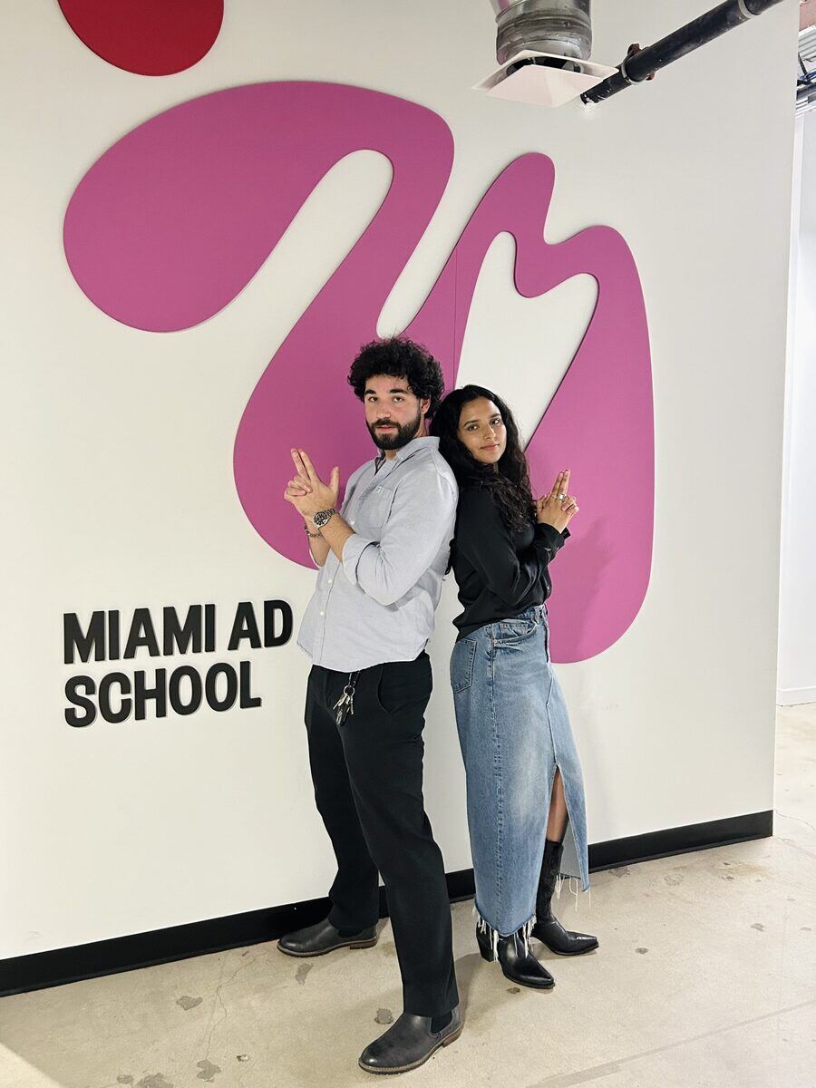 Two individuals standing in front of a wall with the words "Miami Ad School" written on it.